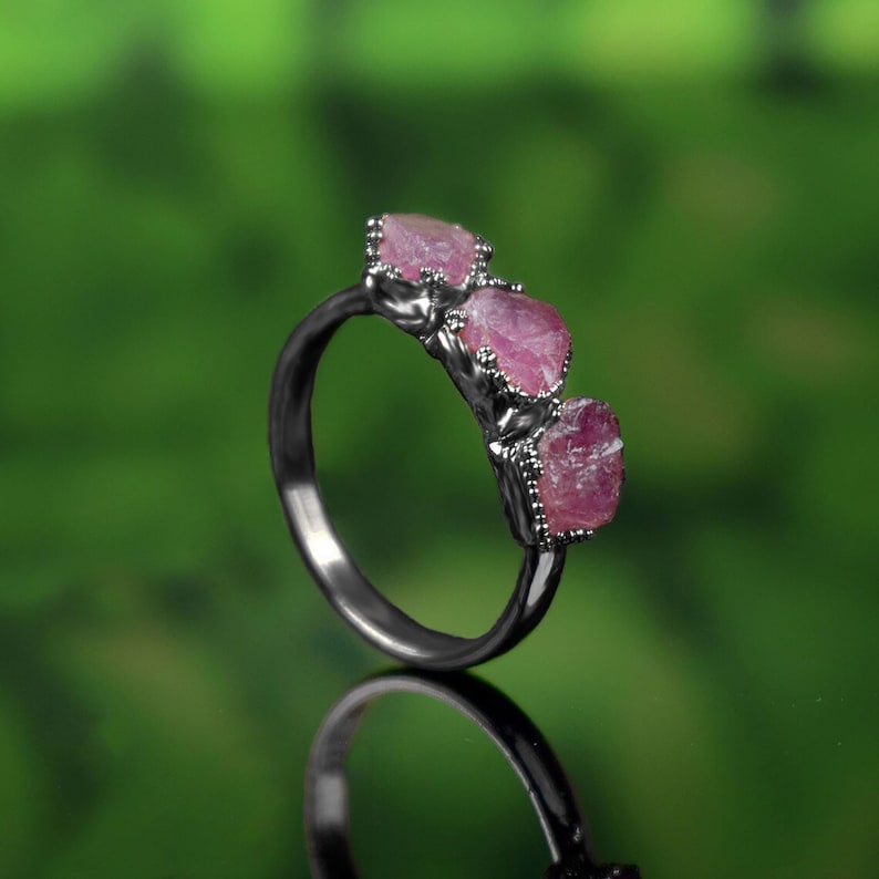 May include: A silver ring with three pink gemstones set in a prong setting. The ring is on a green background.