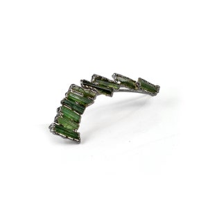 Puede incluir: Un pendiente de plata con un dise&ntilde;o curvo que presenta una fila de piedras preciosas verdes.