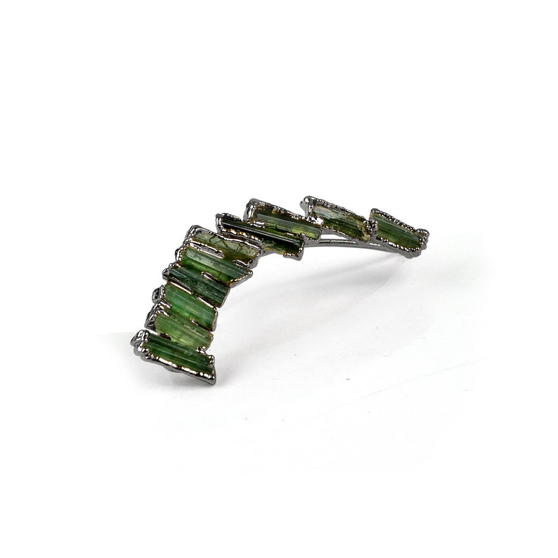 Puede incluir: Un pendiente de plata con un dise&ntilde;o curvo que presenta una fila de piedras preciosas verdes.