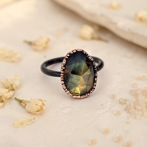 May include: A ring featuring a dark, faceted, iridescent gemstone set in a textured copper-toned bezel. The band of the ring is a smooth, matte black metal. The stone displays shifting hues of green, gold, and grey under the light.