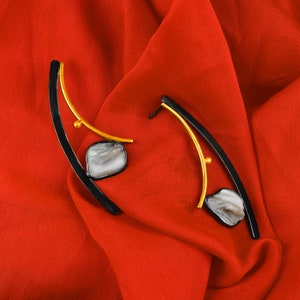 May include: A pair of black and gold earrings with a curved design and a white iridescent stone accent. The earrings are on a red fabric background.