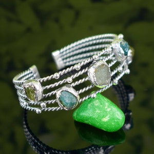 May include: Silver cuff bracelet with five green and blue gemstones set in the twisted silver band. The bracelet is on a green surface.