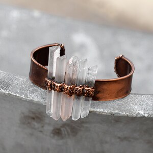 May include: A copper cuff bracelet with clear quartz crystals set in a row. The crystals are arranged in a staggered pattern and are held in place by small copper beads.