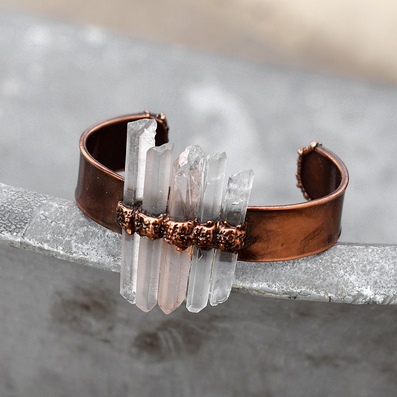 May include: A copper cuff bracelet with clear quartz crystals set in a row. The crystals are arranged in a staggered pattern and are held in place by small copper beads.