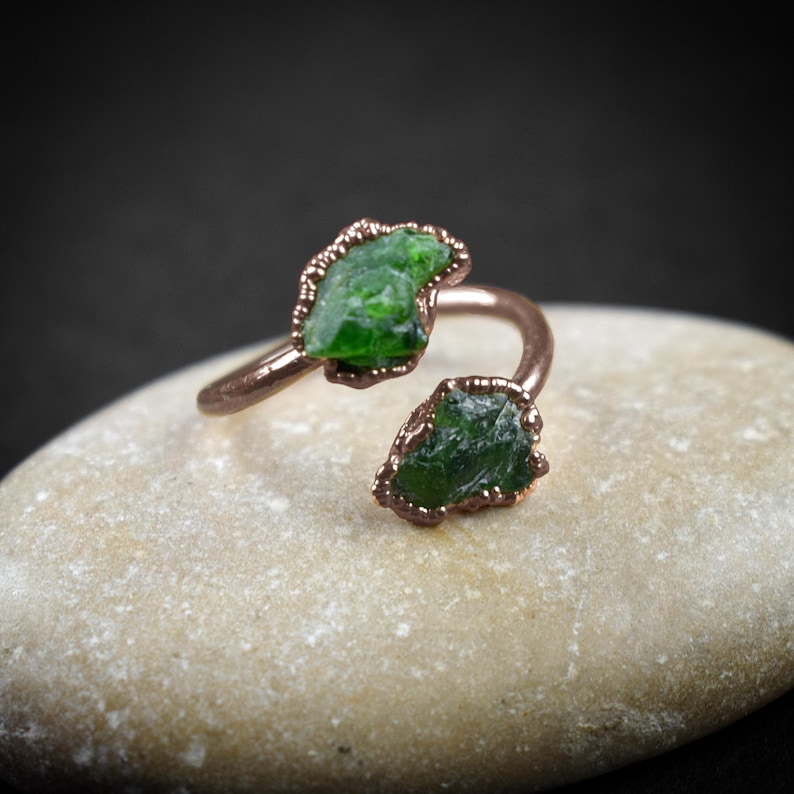 May include: A copper ring with two green gemstone accents. The ring is adjustable and has a unique, twisted design.