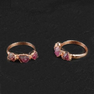 May include: Two gold rings with three pink and red raw ruby stones each. The rings are on a black background.
