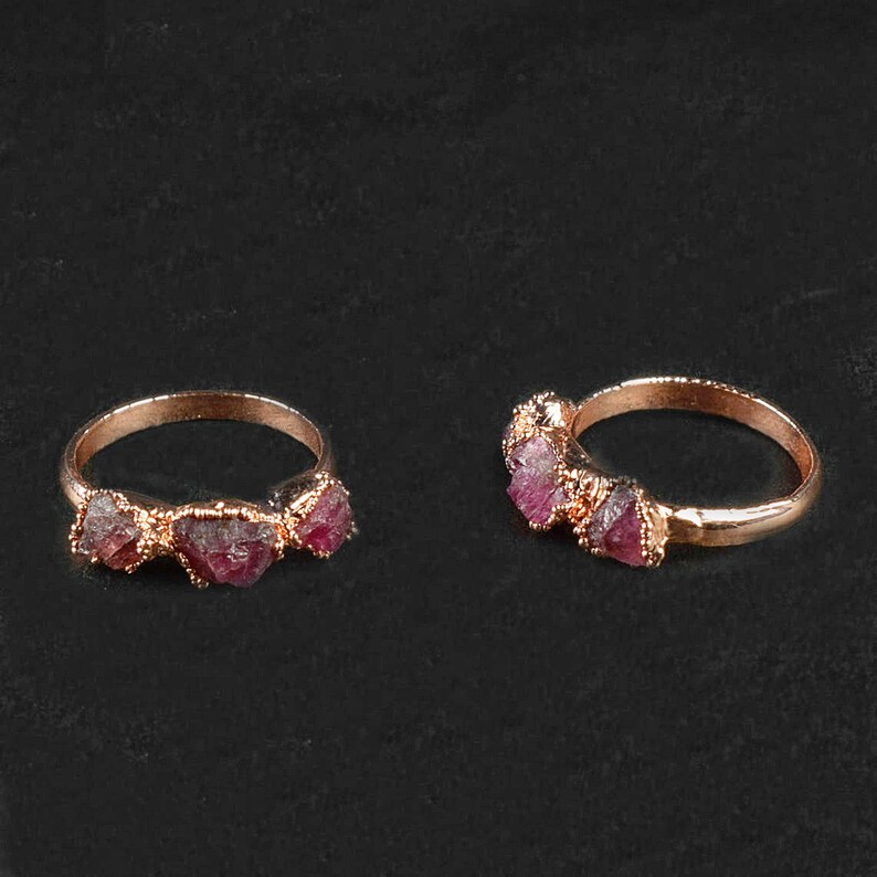 May include: Two gold rings with three pink and red raw ruby stones each. The rings are on a black background.