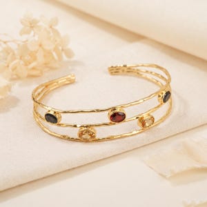 May include: A gold-toned cuff bracelet featuring a double-band design with a hammered texture. The bracelet is accented with four oval-shaped gemstones in shades of deep red, dark black, and light yellow, set securely along the metal bands.