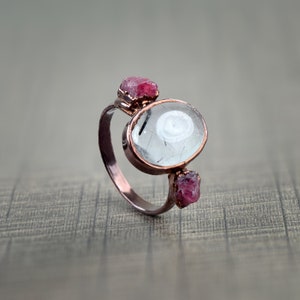 May include: A copper ring with a clear oval-shaped gemstone and two smaller pink gemstones. The ring is on a textured gray surface.