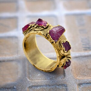 Pode incluir: Um anel de ouro com uma banda texturizada e cinco pedras preciosas vermelhas. O anel est&aacute; colocado sobre uma superf&iacute;cie de azulejos.