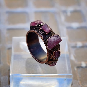 Pode incluir: Um anel de cobre com uma banda de pedras preciosas brutas, cor-de-rosa e roxas engastadas no metal. O anel est&aacute; em um expositor transparente.