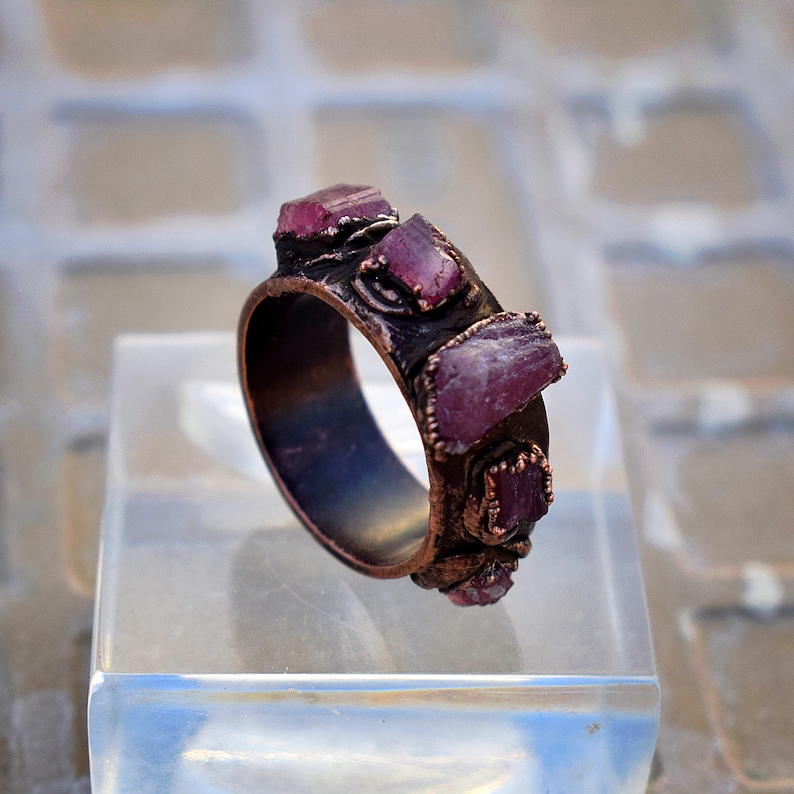 Pode incluir: Um anel de cobre com uma banda de pedras preciosas brutas, cor-de-rosa e roxas engastadas no metal. O anel est&aacute; em um expositor transparente.