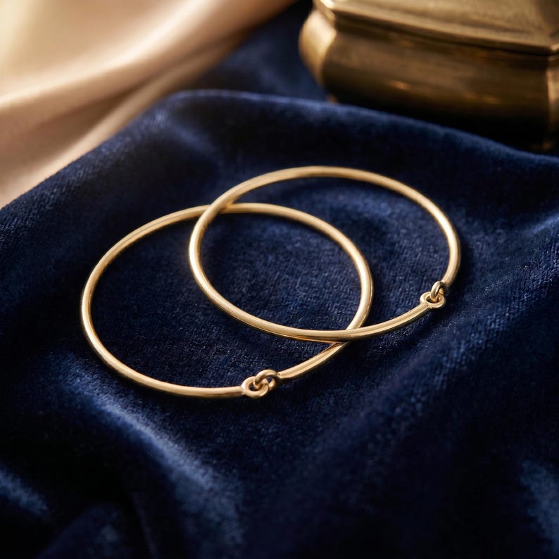 May include: Two thin, circular gold bangles, each featuring a small, simple hook-and-eye clasp mechanism. The bracelets are constructed from smooth, polished gold wire, forming a continuous, minimalist loop when fastened together.