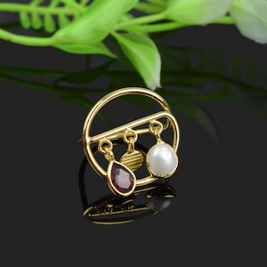Peut inclure: Une bague en or avec un design circulaire et trois breloques pendantes. Les breloques sont une perle blanche, une pierre précieuse rouge et un disque en or.