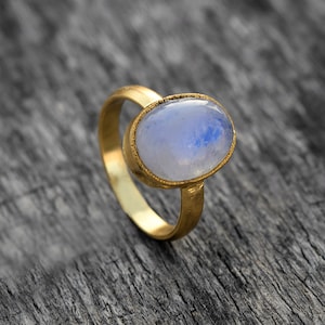 May include: A gold ring featuring a large oval moonstone. The moonstone is a pale blue colour with a subtle iridescent sheen.