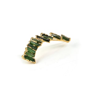Puede incluir: Un pendiente de aro de oro con un dise&ntilde;o curvo que presenta una fila de piedras preciosas verdes.