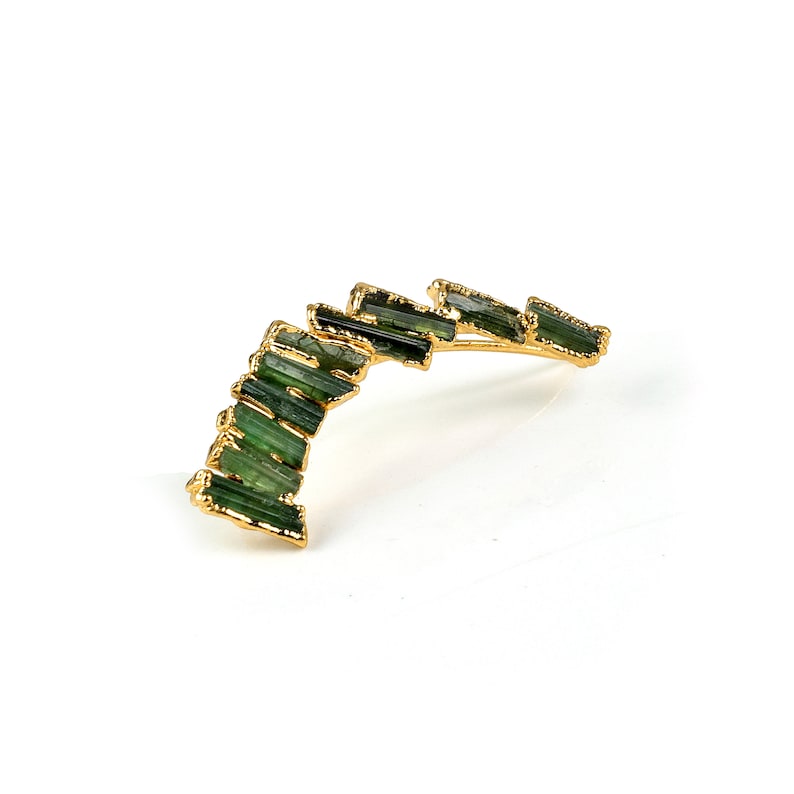 Puede incluir: Un pendiente de aro de oro con un dise&ntilde;o curvo que presenta una fila de piedras preciosas verdes.