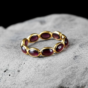 Peut inclure: Une bague dorée ornée de pierres précieuses ovales, d'un violet profond. Les pierres sont disposées en continu. La bague est présentée sur une surface grise texturée, avec un fond sombre. Le design est élégant et attrayant.