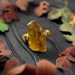 Peut inclure: Une bague en or avec une grande pierre précieuse jaune brute. La bague est posée sur un fond noir avec des feuilles brunes.