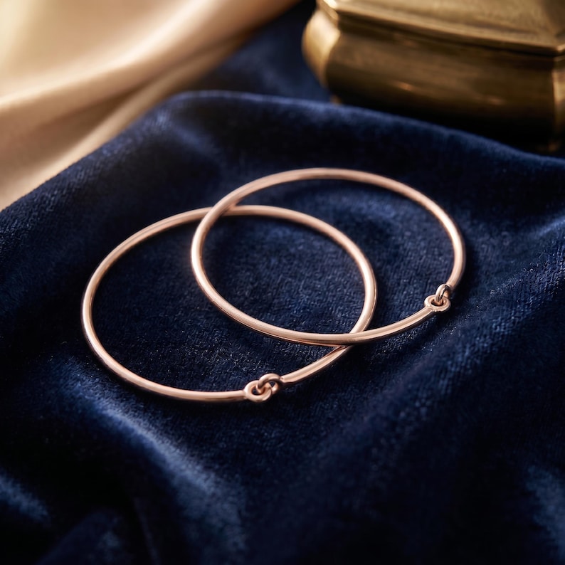 May include: Two thin rose gold bangles, each featuring a simple circular band with a small, integrated loop closure mechanism. The bracelets have a smooth, polished metallic finish and are positioned to overlap slightly, showing their round shape.