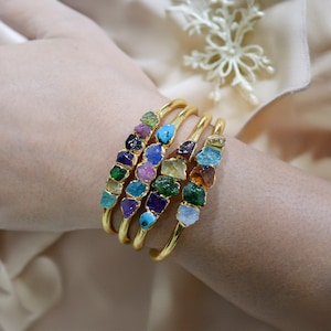 May include: Four gold bangles with a row of colorful, raw gemstones on each. The gemstones are in various shades of green, blue, purple, pink, and orange.
