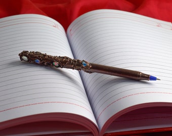 May include: A blank lined notebook with a copper pen with decorative gems on a red fabric background.