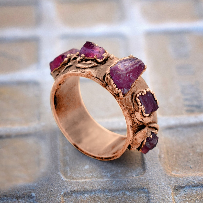 Pode incluir: Um anel de cobre com uma banda texturizada e quatro pedras preciosas cor-de-rosa engastadas na banda.