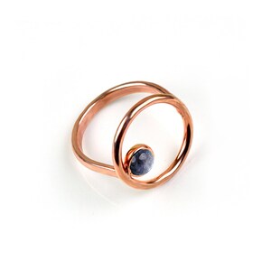 Puede incluir: Un anillo de oro rosa con una sola piedra preciosa redonda gris engastada en un c&iacute;rculo dentro de un c&iacute;rculo m&aacute;s grande.