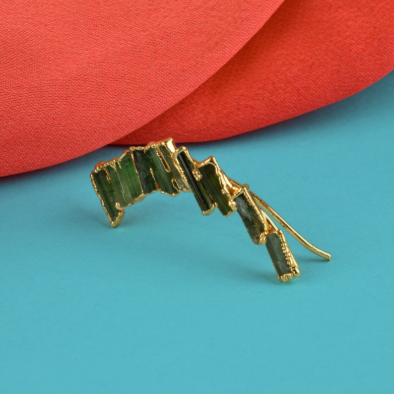 Puede incluir: Pendiente de oro con un dise&ntilde;o curvo adornado con m&uacute;ltiples piedras preciosas verdes.