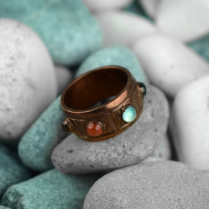 Può includere: Un anello in rame con tre pietre preziose. L'anello ha una fascia testurizzata e presenta una pietra rossa, una blu e una blu chiaro.