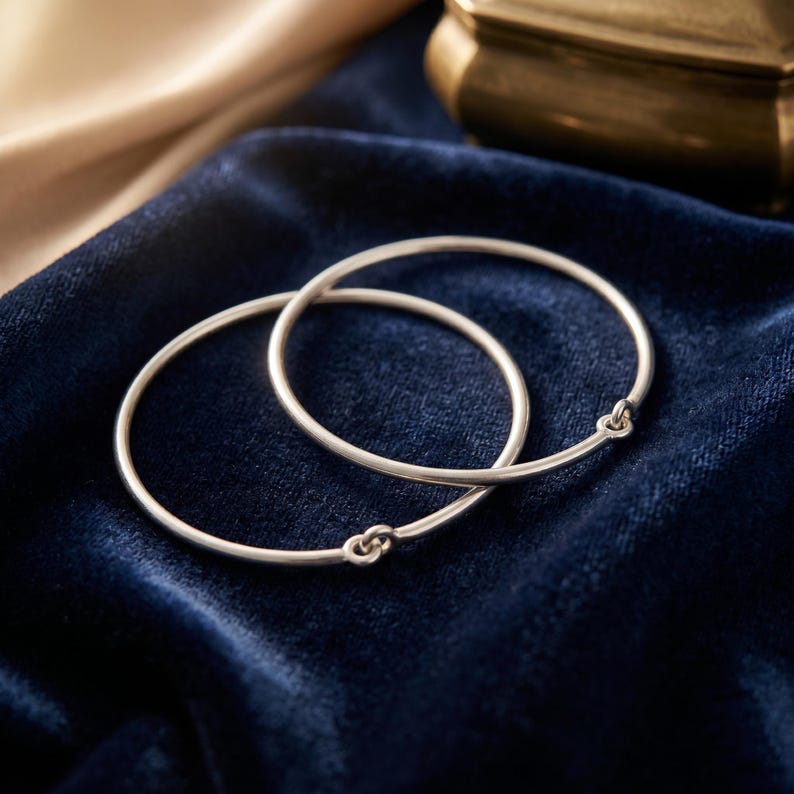 May include: Two thin, circular silver bangles, each featuring a small, integrated loop closure mechanism. The bracelets have a smooth, polished metallic finish and are positioned overlapping one another, showcasing a simple, continuous circular design.