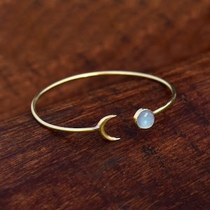 以下が含まれることがあります： 三日月と丸い水色の宝石をあしらった、ゴールドカラーのオープンカフブレスレット。ブレスレットは濃い木目の背景に配置され、ジュエリーのデザインを際立たせています。