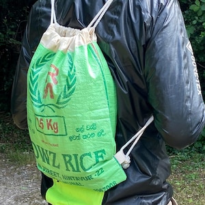May include: A green and white drawstring backpack made from a rice sack. The bag features text and graphics, including "15 Kg" and "AWL RICE". The bag is carried by a person wearing a black jacket and a cap.