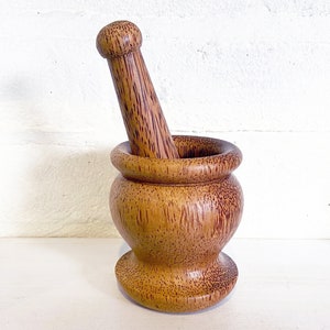 May include: A wooden mortar and pestle set made from coconut wood. The mortar is brown with a reddish hue and the pestle is a light brown color.
