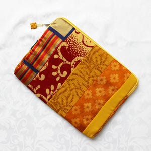 May include: A rectangular laptop sleeve featuring a patchwork design of various fabrics in shades of red, orange, and yellow. The surface displays patterns including stripes, floral motifs, and geometric shapes, finished with a yellow zip closure.