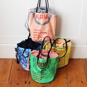 May include: Several tote bags made from recycled rice sacks. The bags are in shades of peach, green, and yellow, with black handles. The bags have printed text and graphics, including "RRM" and "USDA RICE". The bags are filled with fabric.