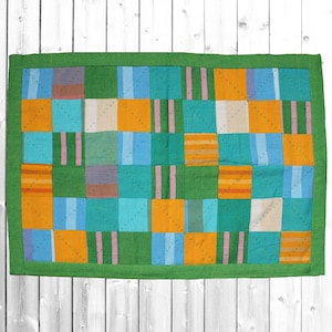 Könnte beinhalten: Eine Patchwork-Decke mit grünem Rand. Die Decke besteht aus Quadraten in verschiedenen Farben, darunter Orange, Blau, Grün und Braun.