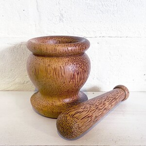May include: A wooden mortar and pestle set made from natural wood. The mortar is brown with a reddish hue and has a round, wide base. The pestle is a long, cylindrical shape with a rounded end.