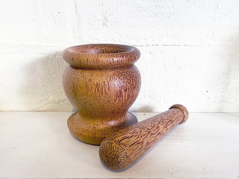 May include: A wooden mortar and pestle set made from natural wood. The mortar is brown with a reddish hue and has a round, wide base. The pestle is a long, cylindrical shape with a rounded end.
