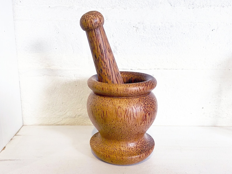 May include: A wooden mortar and pestle set made from coconut wood. The mortar is brown with a reddish hue and the pestle is a light brown color.