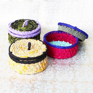 May include: Four handmade woven storage boxes in various colours. One box is yellow and black, another is green and purple, and a third is pink and blue. The fourth box is open, revealing a blue interior. The boxes are round and have lids.
