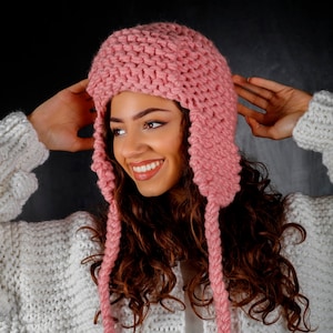 May include: A pink knitted hat with long ear flaps. The hat is made of a chunky yarn and has a simple, classic design.