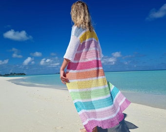 Op de afbeelding: Een kleurrijke, open geweven, regenboog gestreepte cover-up. De cover-up heeft korte mouwen en een geschulpte zoom. De cover-up wordt gedragen op een strand met wit zand en turquoise water onder een helderblauwe lucht.