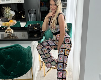 Op de afbeelding: Een vrouw draagt een zwart en veelkleurig gehaakt jumpsuit met een vierkant patroon. Ze zit op een gouden stoel in een keuken.