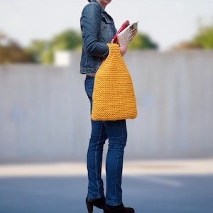 Könnte beinhalten: Eine Frau trägt eine gelbe Stricktasche. Die Tasche ist aus dickem, strukturiertem Garn gefertigt und hat eine einfache, rechteckige Form.