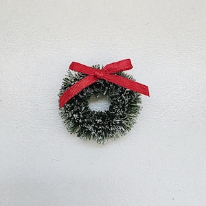 May include: A small Christmas wreath featuring a red ribbon bow. The wreath is made of green, snow-covered pine needles. The bow is a vibrant red and is tied at the top of the wreath. A festive decoration for the holiday season.