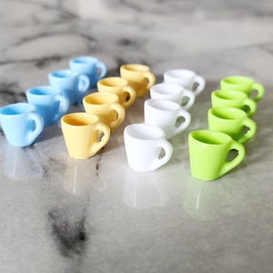 May include: A collection of miniature ceramic mugs in various colors, including light blue, yellow, white, and green. The mugs are arranged in a staggered pattern on a white surface.