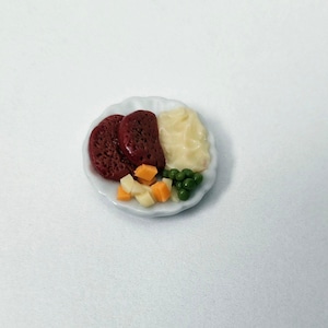 Peut inclure: Une assiette miniature en céramique présentant un repas composé de deux steaks hachés rouge foncé, une portion de purée de pommes de terre blanche crémeuse, des dés de légumes orange et blancs, et quelques petits pois verts sur un fond blanc.