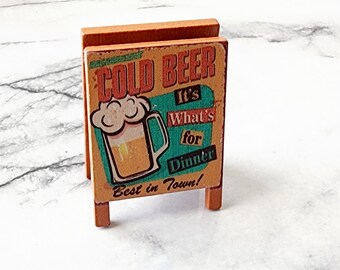 May include: A miniature wooden sign with a vintage design advertising "Cold Beer" with a cartoon image of a glass of beer and the text "It's What's for Dinner! Best in Town!"