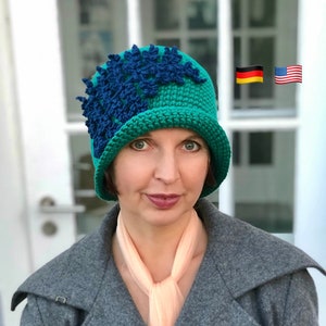 May include: A teal blue cloche hat with a blue crocheted flower design. The hat is worn by a person with a peach colored scarf and a gray coat.